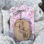 Wooden keychain with 'Be You' text on a pink and white patterned card, placed on textured fabric.