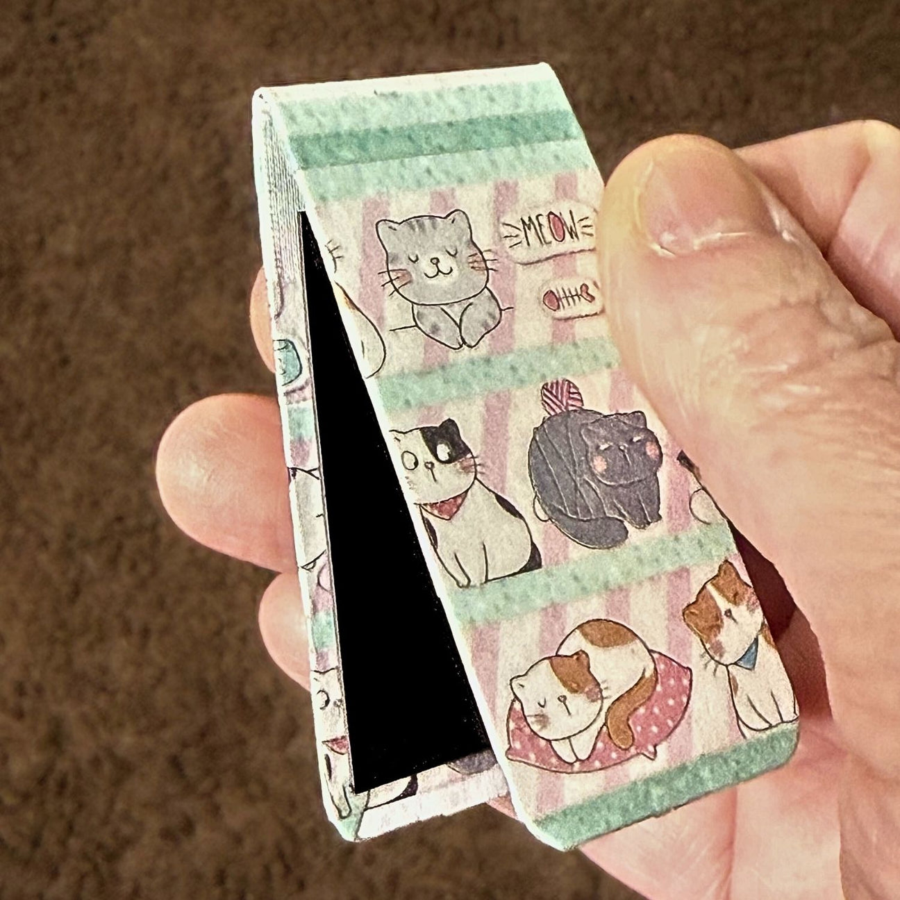 Magnetic bookmark with cat illustrations held by a hand against a brown background