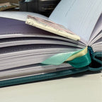 Open book with a green ribbon and a profile view of a magnetic bookmark on a light surface.