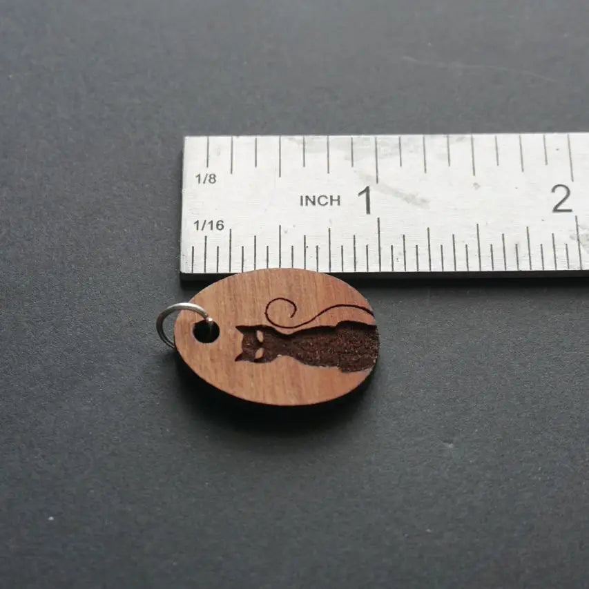 Wooden pendant with engraved cat design next to a ruler on a dark surface