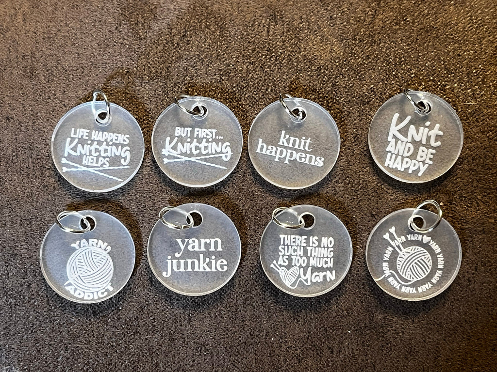 A set of nine clear acrylic stitch markers with engraved knitting-related phrases, displayed on a textured background.