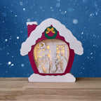 A red paper cut house scene lit up with a family Christmas scene inside sitting on a table against a blue snowy background.